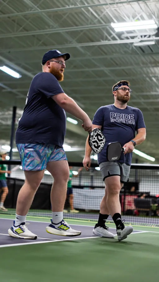 Two pickleball players walk together on an indoor court, each holding a paddle and wearing athletic clothing including shorts, t-shirts with "Pride Sports USA" text, and sports shoes; one player also has a headband and knee support.