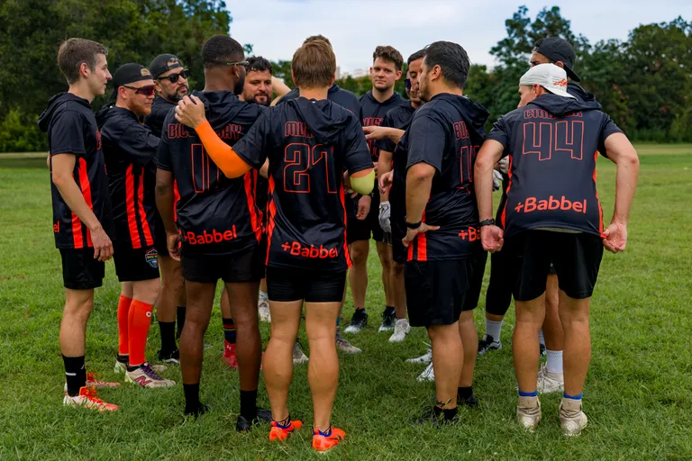 ATX Heat (Orange) players huddle on a grassy park field during practice for Gay Bowl 2025, listening and gesturing before a drill. They wear black jerseys with orange accents, numbers, and sponsor text, with shorts and cleats; some athletes also have caps, headbands, or arm/leg sleeves, and trees frame the background.