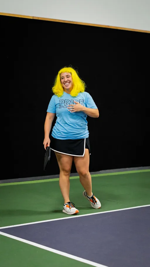 A pickleball player wearing a bright yellow wig, a blue "Pride Sports" shirt, black shorts, and athletic shoes smiles on an indoor court while holding a paddle.