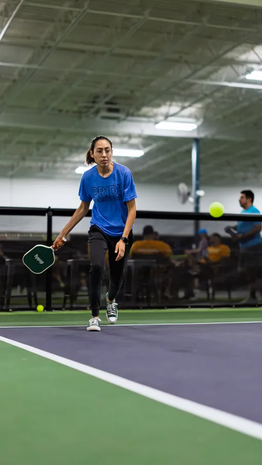 A pickleball player wearing a blue "PRIDE" t-shirt and black athletic pants is in motion on an indoor pickleball court, preparing to hit a ball with a paddle. There are other participants and spectators visible in the background.