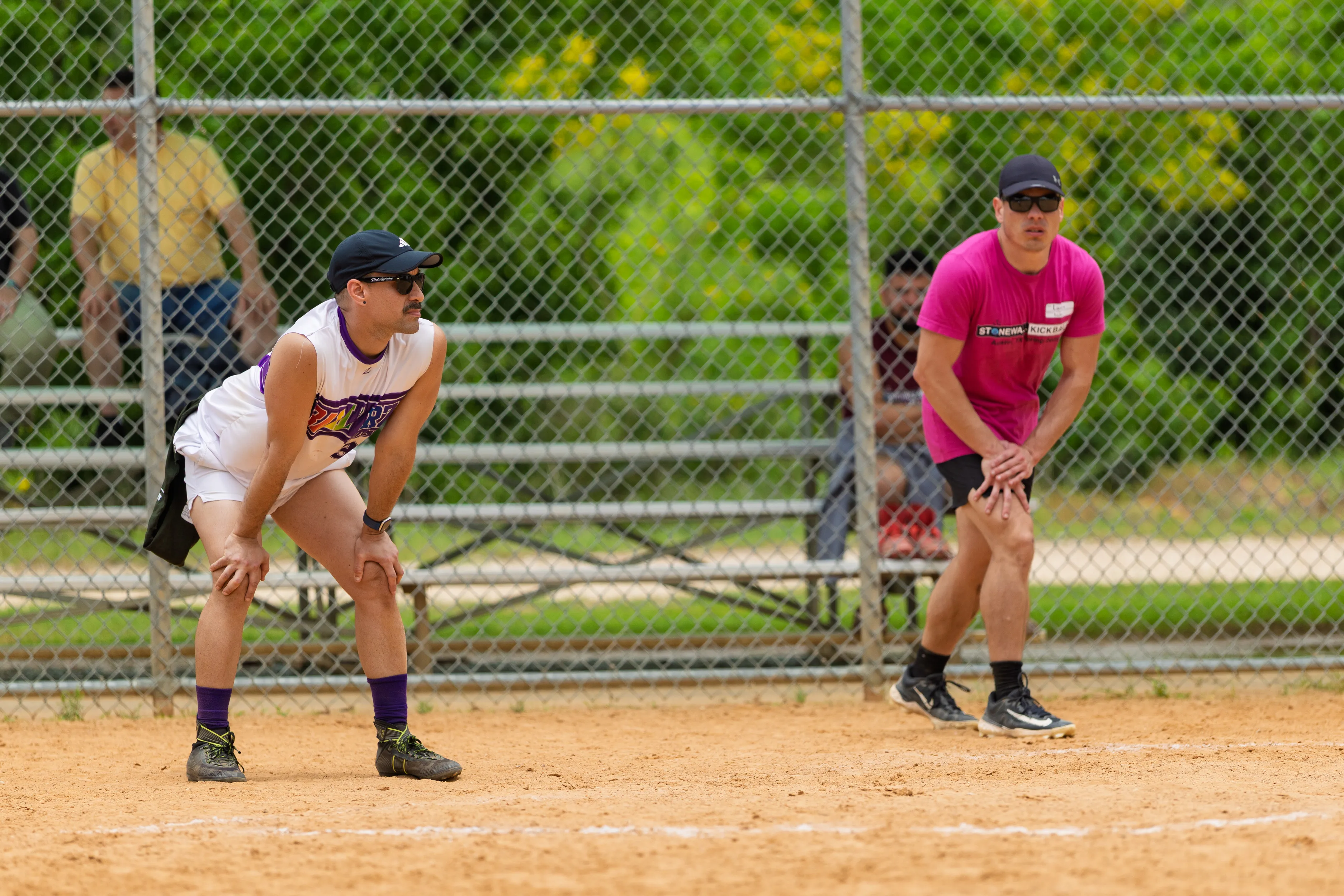 Two players on a kickball infield hold a defensive ready stance with hands on knees, watching for the next play. One wears a white sleeveless jersey with purple trim, black cap, sunglasses, purple socks, and athletic shoes; the other wears a bright pink shirt with a name tag, black shorts, black cap, and sunglasses, with a chain-link fence and bleachers in the background.