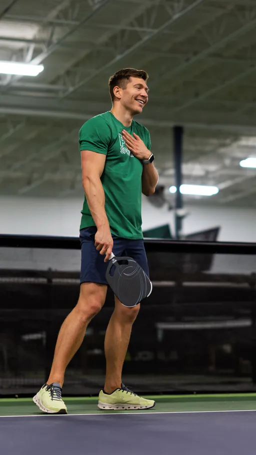 A pickleball player is standing on an indoor court, holding a paddle and smiling, wearing athletic shoes, navy shorts, and a green t-shirt. The background features high ceilings and bright lighting typical of an indoor sports facility.