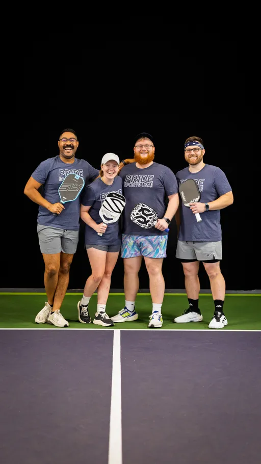 Four pickleball players stand together on a pickleball court, each holding a paddle and wearing matching "Pride Sports USA" shirts, athletic shorts, and sneakers, with two players wearing hats or visors. They are smiling and appear relaxed and happy, posing as a team before or after a match.