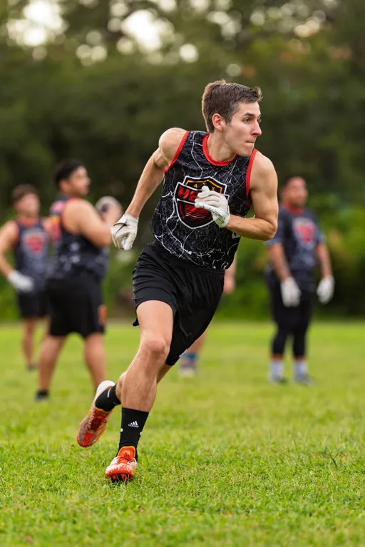 An ATX Heat athlete sprints on a grassy field during flag football practice, wearing a sleeveless black team jersey with red trim and crest, black shorts, white gloves, and orange cleats. Teammates in matching jerseys and gloves practice in the background with trees lining the field.