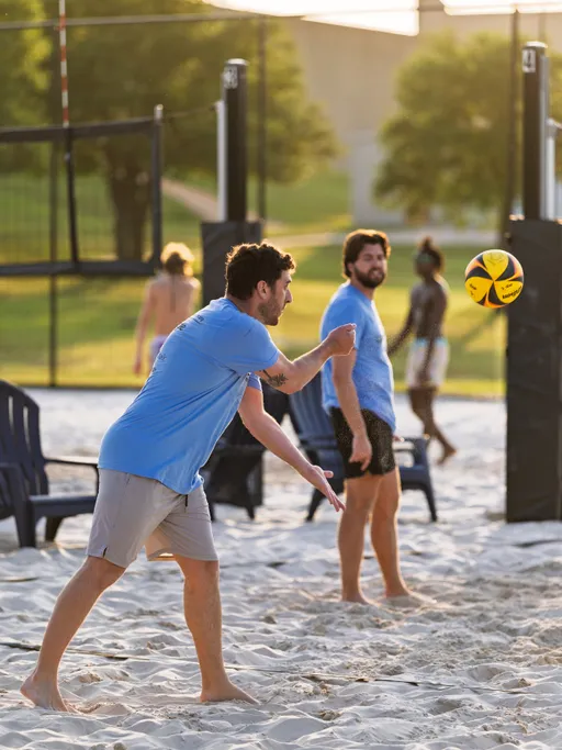 A volleyball player in a light blue shirt and light shorts is serving a ball on a sand court, while another player in a matching shirt stands nearby; outdoor volleyball nets and several people in the background are visible.