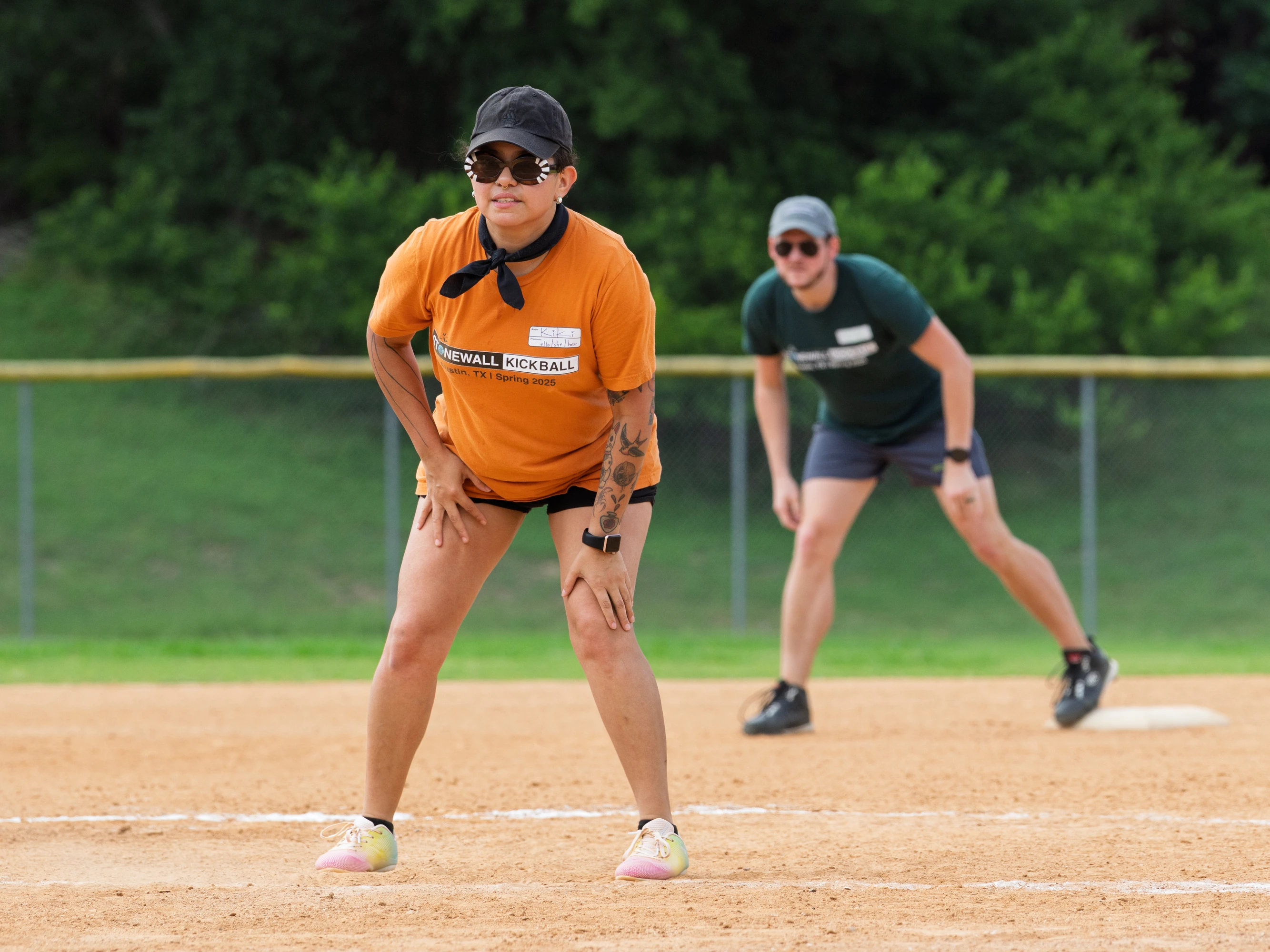 Two kickball players are on a dirt field, with one in the foreground wearing an orange Stonewall Kickball shirt, athletic shoes, a black cap, sunglasses, and a bandana, poised and focused on the game, while a second player in a green shirt stands ready in the background near a base.
