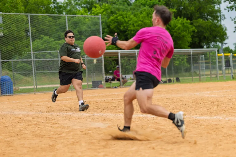 A player in a pink shirt and black shorts runs toward a base on a sandy kickball field as another player in a dark green shirt and black shorts throws or pitches a red kickball toward them.