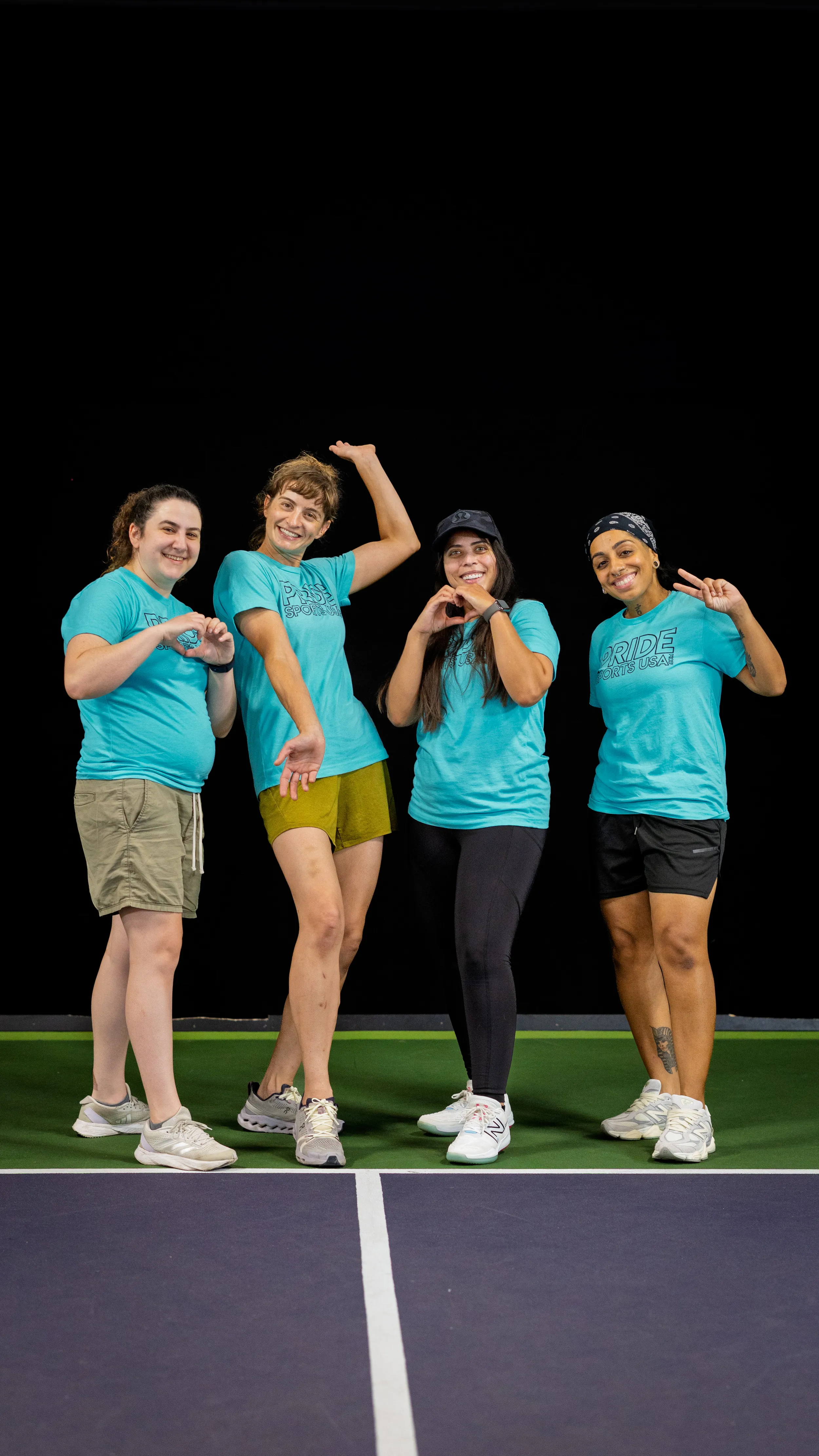 Four pickleball players in matching teal “PRIDE Pickleball” shirts pose together on an indoor court, smiling and striking playful gestures like peace signs and pointing. They wear athletic shorts or leggings and sneakers, with accessories including a cap and a headband; court lines are visible in the foreground against a dark backdrop.