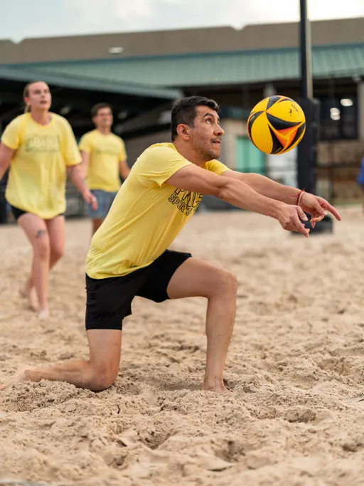 A player in a yellow shirt and black shorts is kneeling in the sand, extending their arms to bump a volleyball, while other athletes in yellow shirts move behind them on an outdoor sand court.