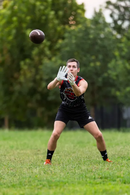 On a grassy park field, an ATX Heat (Orange) player in a black sleeveless ATX Heat jersey, black shorts, white receiver gloves, and orange cleats focuses on a football in midair with hands open to catch it. Trees blur in the background during practice for Gay Bowl 2025.