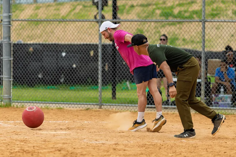 Two kickball players are running on a dirt field, with one in a bright pink shirt and white hat and the other in a dark green shirt and black cap; a red kickball rolls across the ground while spectators watch from behind the chain-link fence.