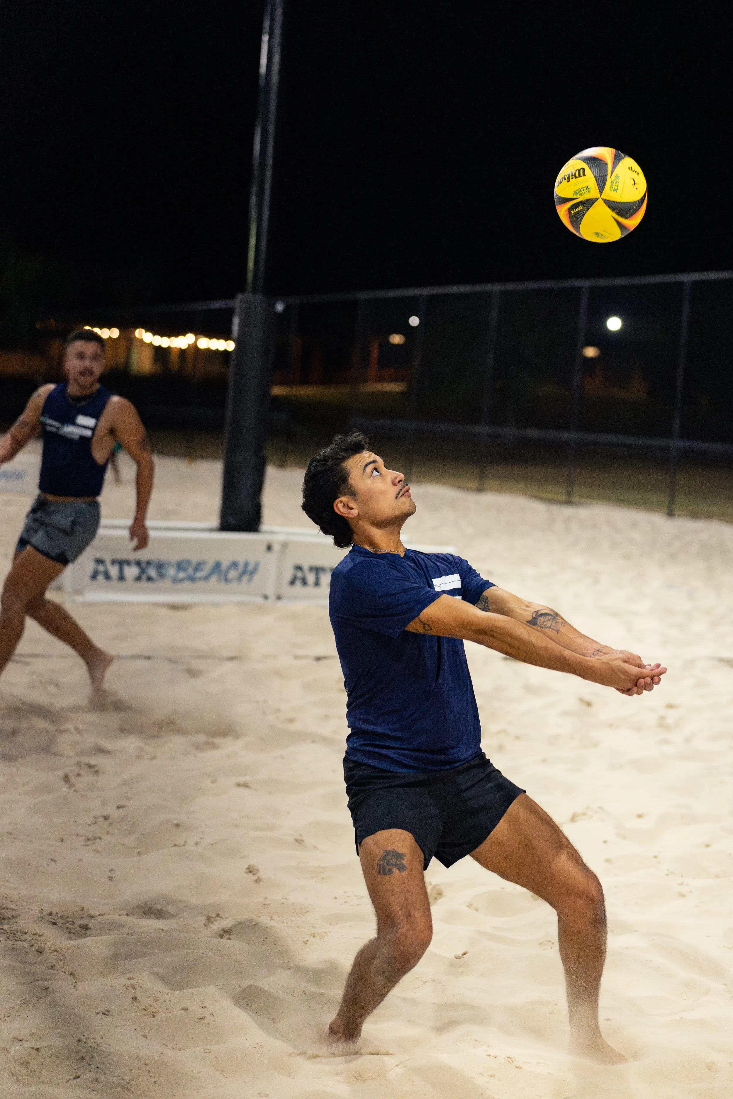 On a lit sand volleyball court at night, a player in a navy shirt and black shorts prepares to bump a yellow-and-blue volleyball, arms together and eyes up. A teammate in a matching top moves behind near the net and an “ATX BEACH” barrier, with fencing and park lights in the background.