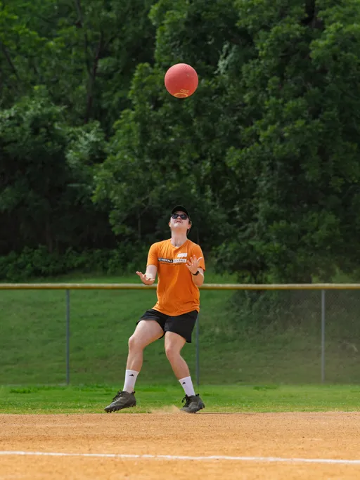 A kickball player wearing an orange shirt, black shorts, white socks, sunglasses, and a black cap prepares to catch a red kickball on a dirt field with green trees in the background.