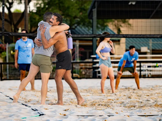 Two volleyball players in athletic clothing are hugging on a sandy court, with one holding a water bottle; other players stand nearby, some preparing for the next play. The scene takes place outdoors with a volleyball net and benches visible in the background.