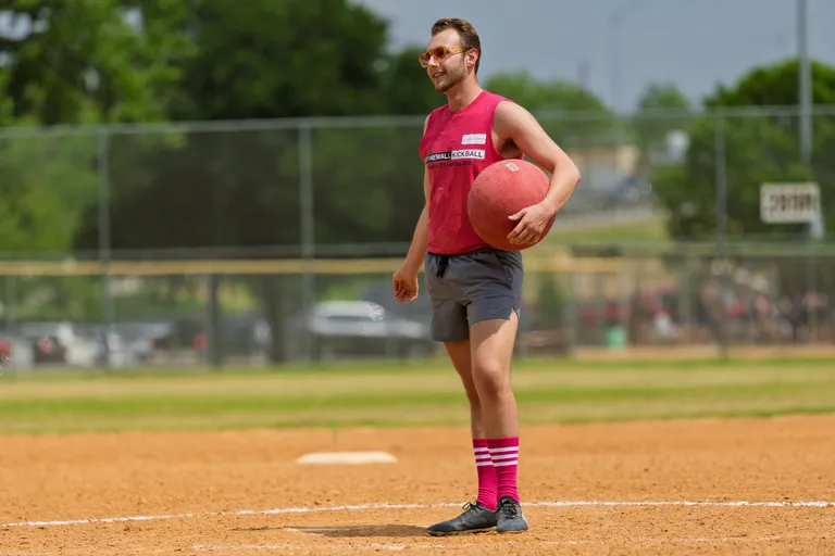 A player stands on a kickball field holding a red kickball, wearing a sleeveless team shirt, shorts, sunglasses, and knee-high pink striped socks, with bases and a fence visible in the background.