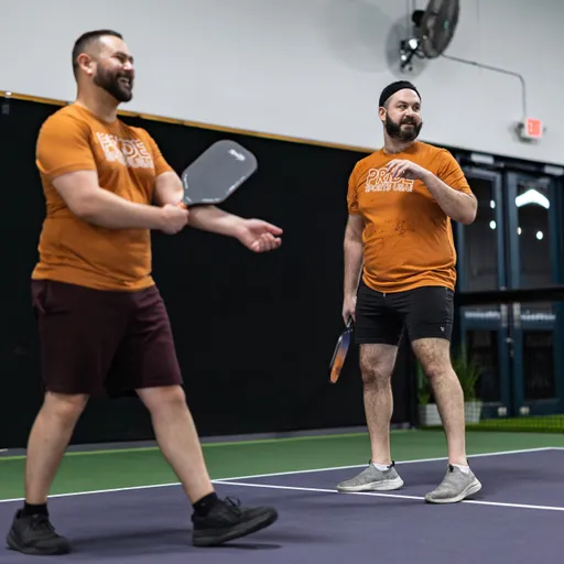 Two pickleball players wearing matching orange "Pride Pickleball" shirts, shorts, and sneakers are on an indoor court; one player is in motion and smiling with a paddle in hand, while the other stands nearby, also holding a paddle, as they prepare for the next play.