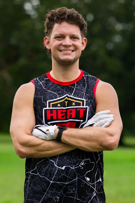 On a grassy practice field, an ATX Heat player stands with arms crossed, wearing a black sleeveless jersey with red trim that reads “ATX HEAT” over a map-style pattern and white receiver gloves. Trees blur in the background as the team preps for Gay Bowl 2025 flag football.