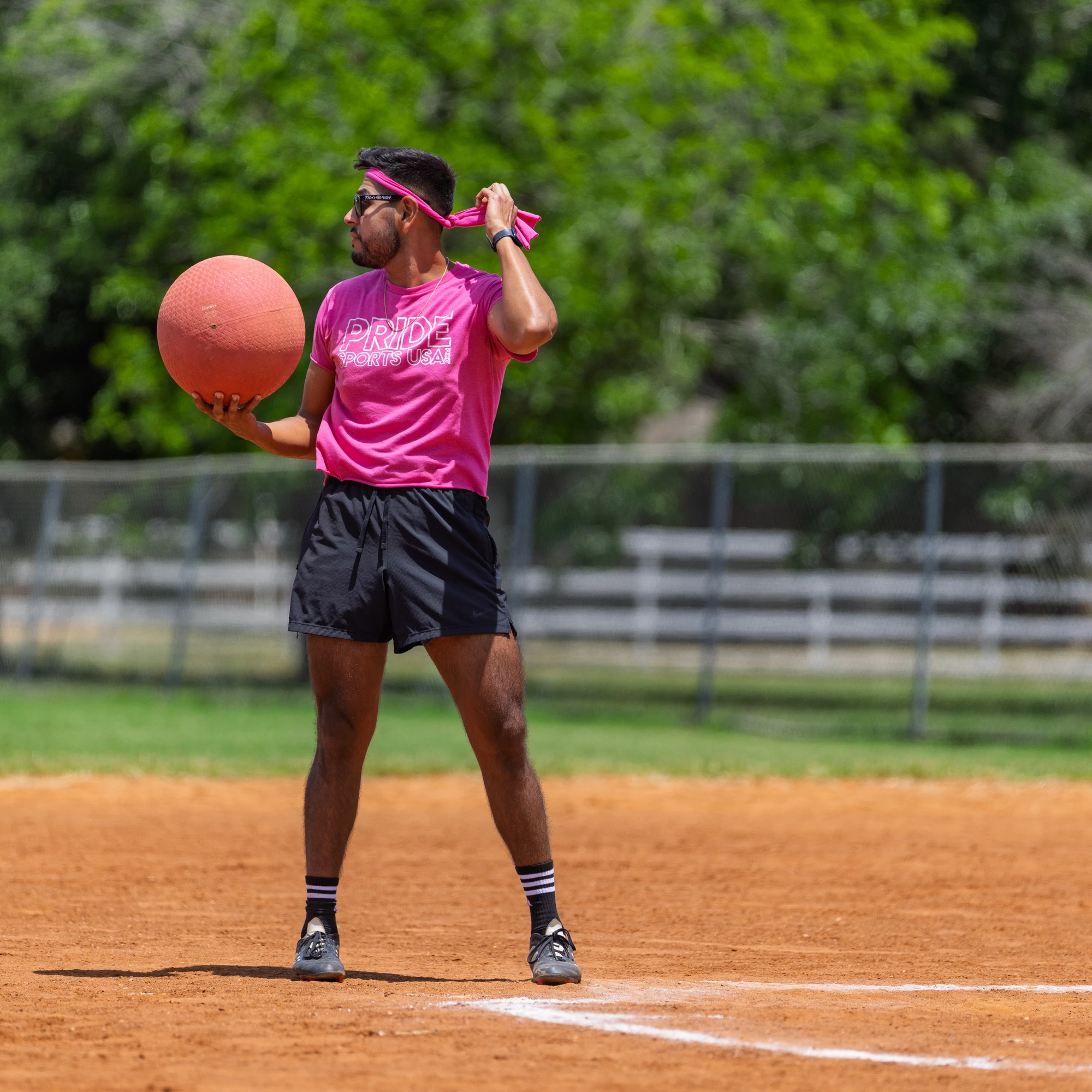 A kickball player in a pink shirt, black shorts, and a matching pink headband prepares to throw a kickball on a sunny outdoor field. The athlete stands near the pitcher's mound, looking toward their target with teammates and trees in the background.