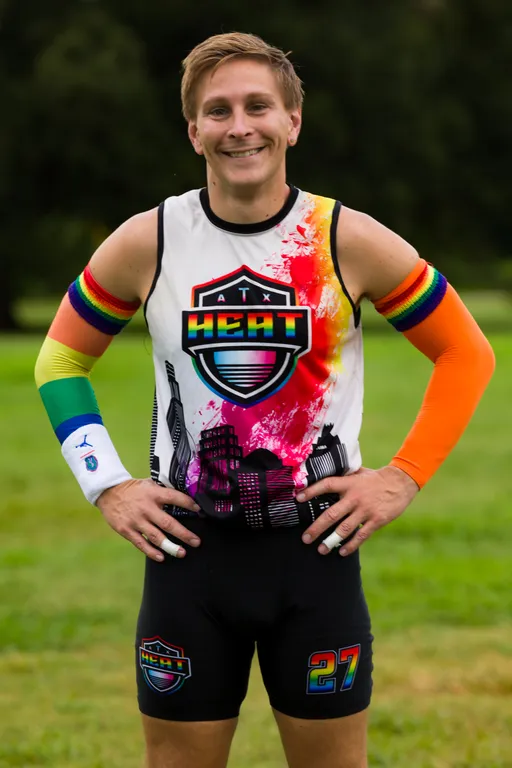 An ATX Heat player poses on a grassy practice field during Gay Bowl 2025 prep, hands on hips and smiling. The athlete wears a white sleeveless jersey with the ATX Heat logo and colorful paint-splash skyline, rainbow arm sleeves (one mostly orange), a white wristband, and black shorts featuring the team crest and number 27.