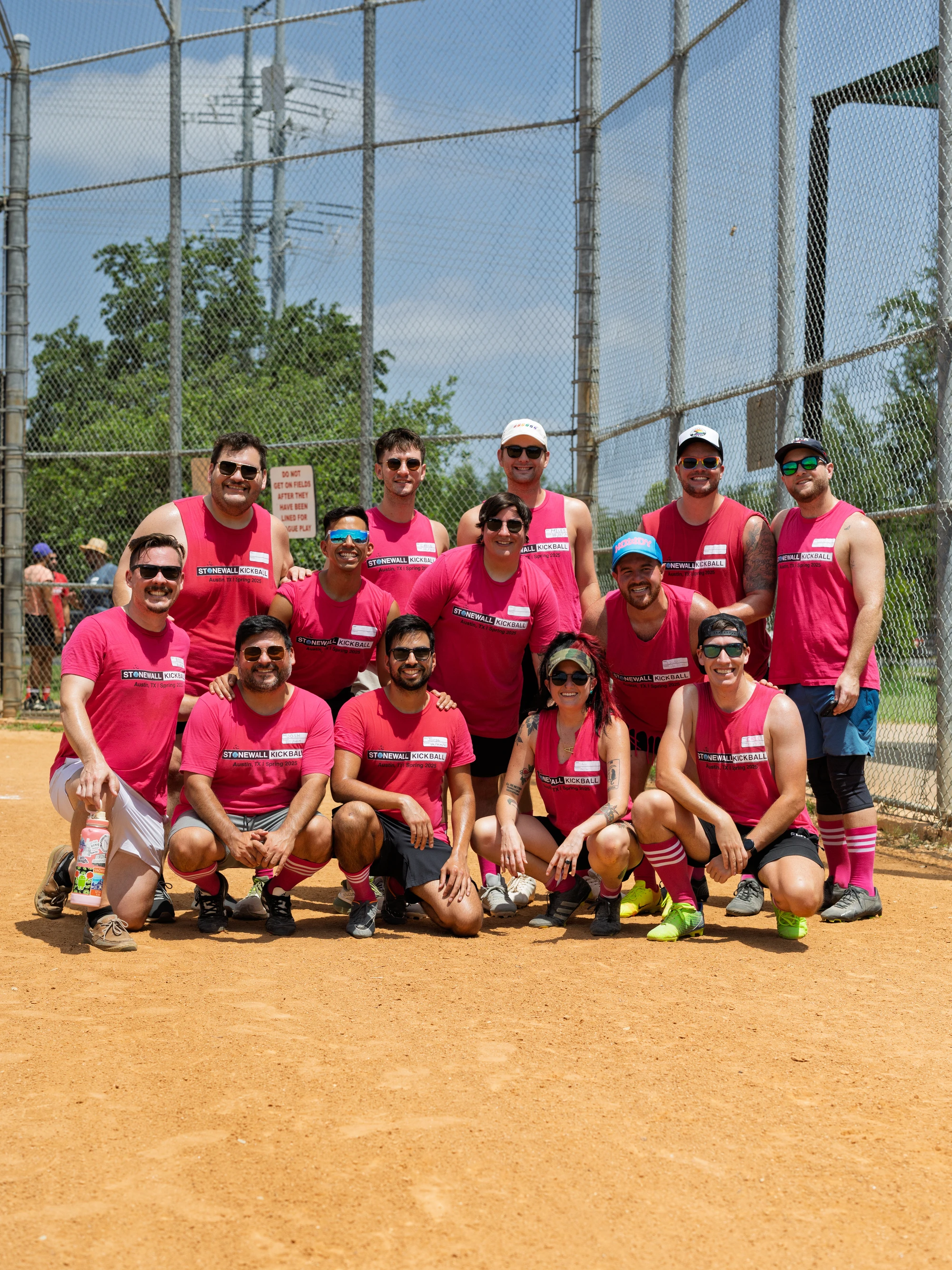 A group of kickball players wearing matching bright pink Stonewall Kickball jerseys pose together on a dirt kickball field, some kneeling in front and others standing behind; several players are wearing sunglasses, hats, or colorful knee-high socks, and a fence and trees are visible in the background.