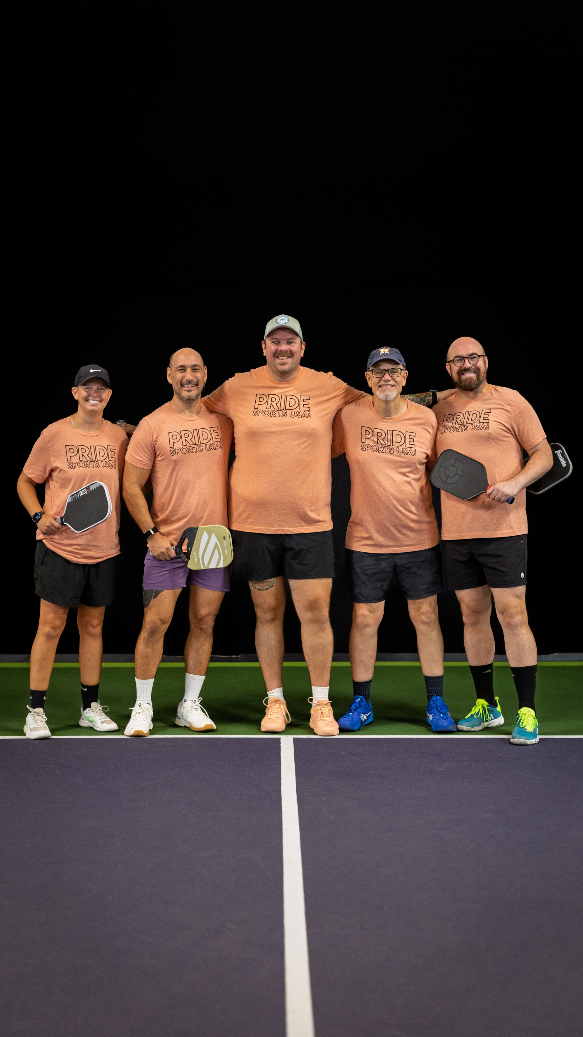 Five pickleball players stand arm in arm on an indoor court, each wearing matching "Pride Pickleball" t-shirts, shorts, and athletic shoes, while holding their pickleball paddles and smiling towards the camera.