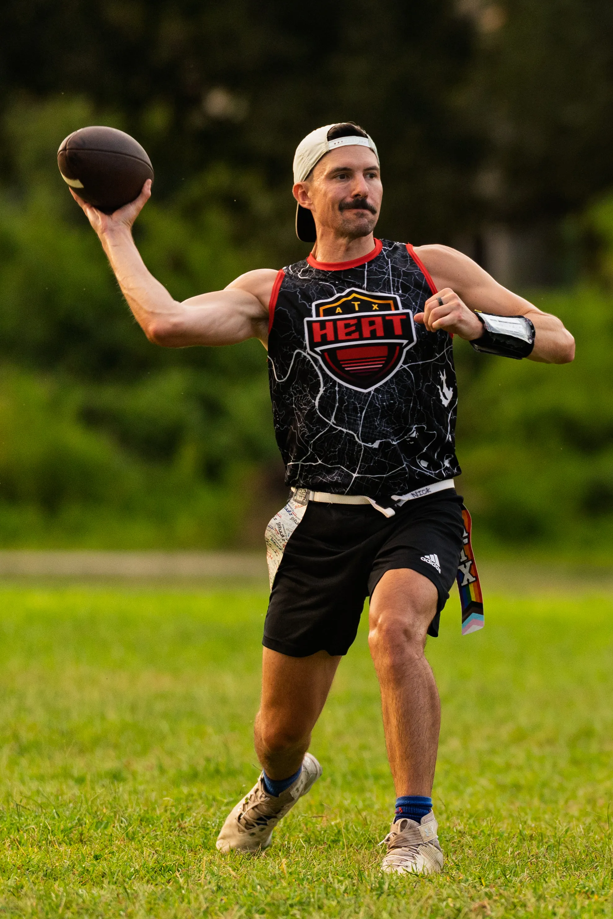 On a grassy field, an ATX Heat (Orange) flag football player winds up to throw a pass, wearing a black sleeveless jersey with the Heat logo, black shorts, cleats, a backward white cap, a wrist play band, and a rainbow-striped flag belt; trees blur in the background during practice for Gay Bowl 2025.