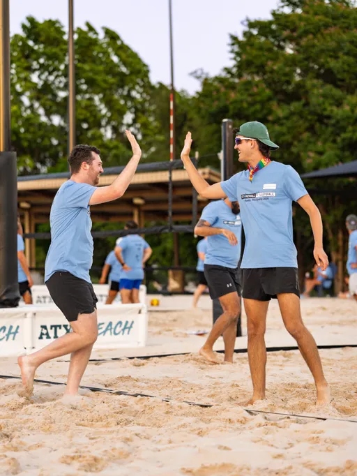 Two volleyball players wearing matching blue team shirts and shorts are giving each other a high five on a sand court during a game, with other players and volleyball equipment visible in the background.