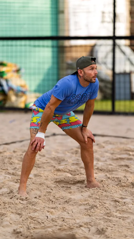 A volleyball player is standing in a ready position on a sand court, wearing a blue "Pride Sports" shirt, colorful shorts, a backwards cap, and a wrist wrap, appearing focused on the game.