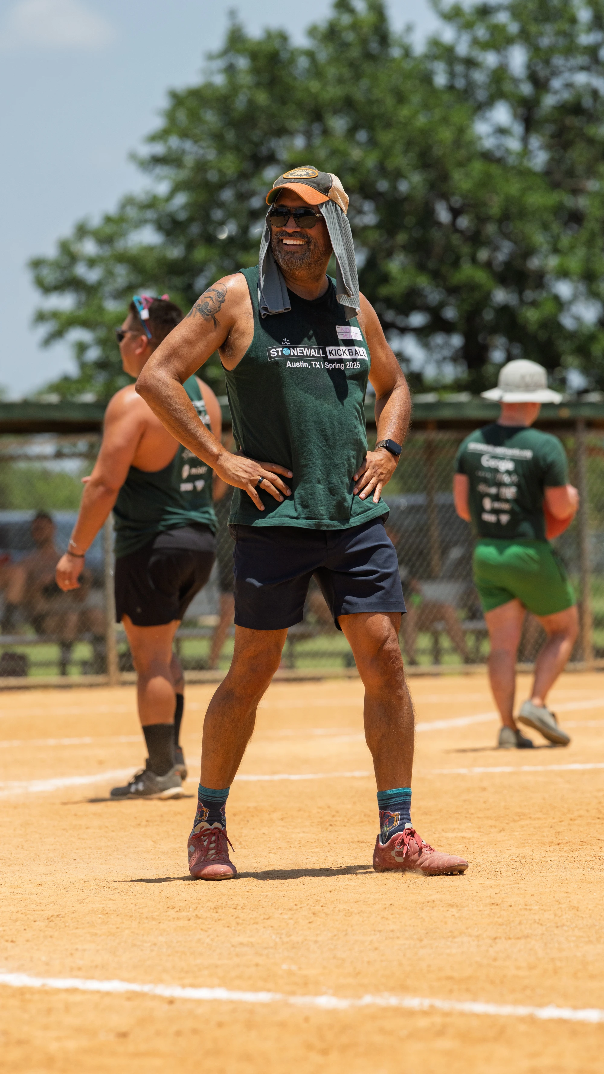 A kickball player stands on the field with hands on hips, wearing a sleeveless team shirt, shorts, sneakers, sunglasses, and a cap with a cloth draped for sun protection; other players and a fence are visible in the background.