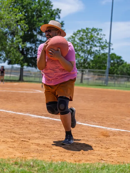 A kickball player in a straw hat, sunglasses, pink shirt, brown shorts, and knee pads sprints across a dirt field while holding a red kickball, with green trees and a fence visible in the background.